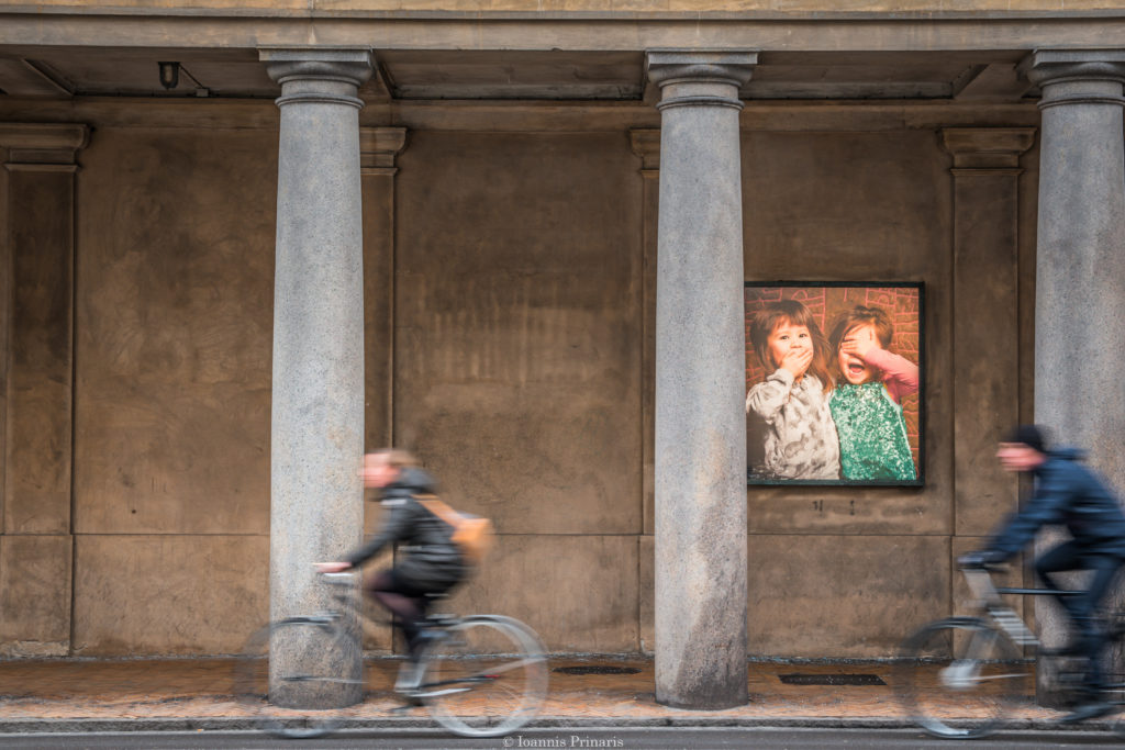 Two people on bicycles in front of a wall where there is a picture of two kids smiling.